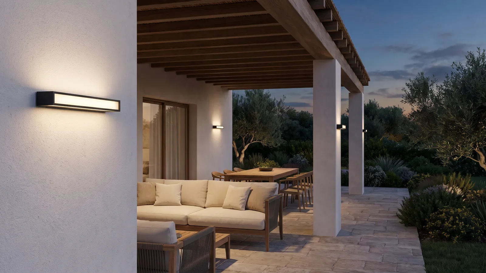 Terraza cubierta mediterránea al anochecer con apliques negros de barra horizontal y difusor frontal opal sobre muros blancos, pérgola de madera, sofá y mesa de exterior, y jardín.